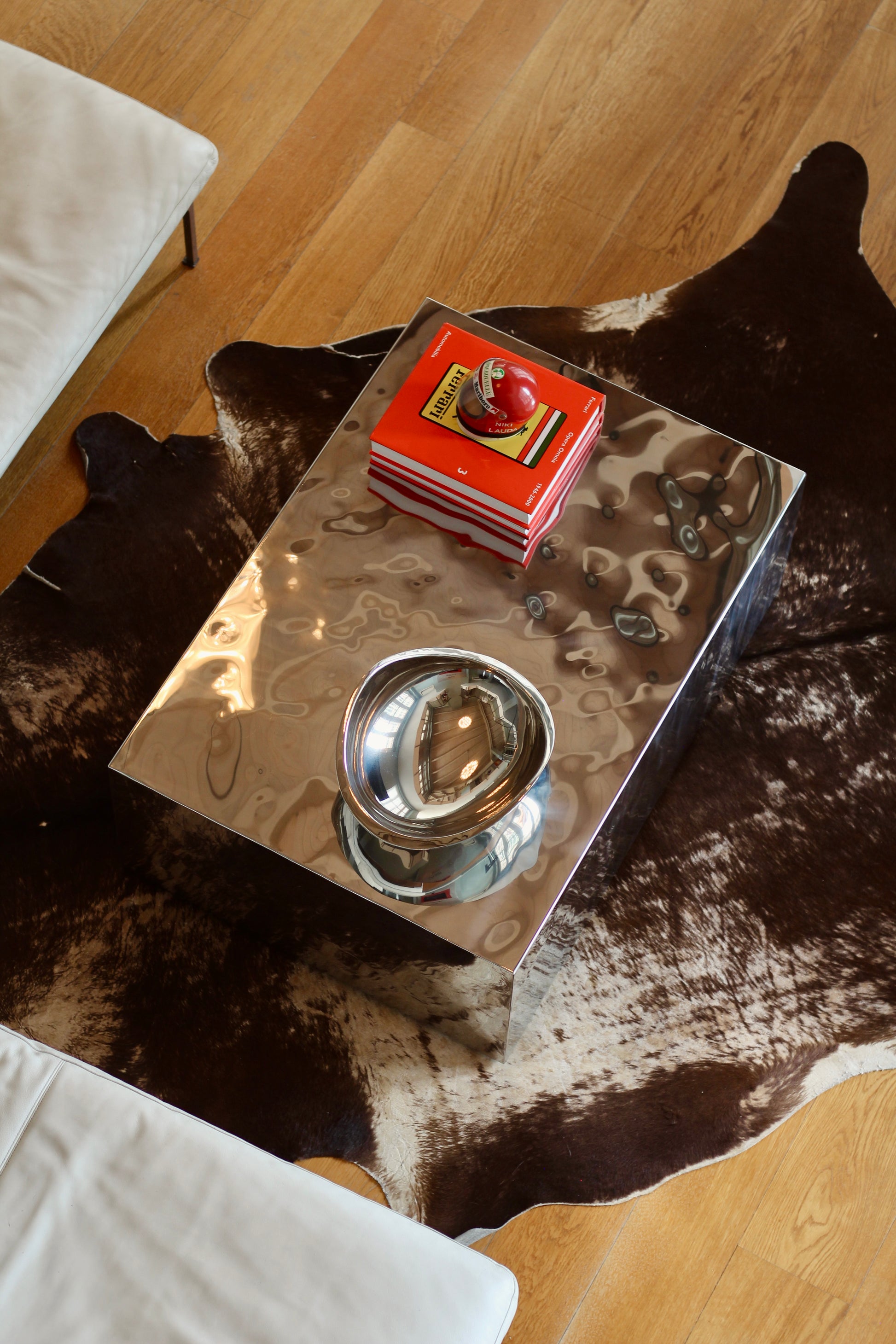 A rectangular polished chrome coffee table with a matching chrome bowl, styled with a red book on a cowhide rug over a light wood floor.