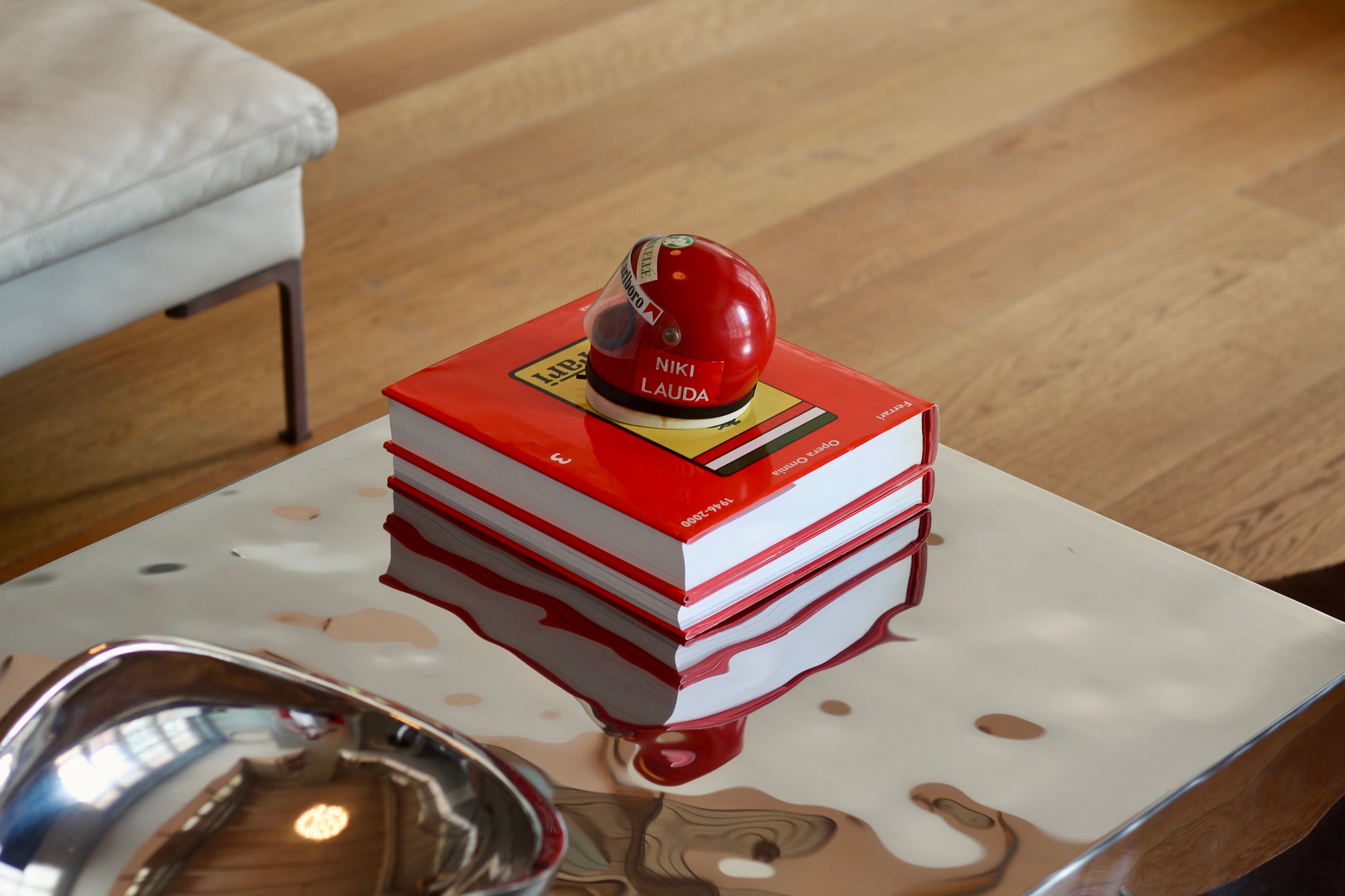 A stack of three red and white hardcover books with a glossy red spherical object on top, placed on a reflective white table in a modern interior setting with a light wood floor.