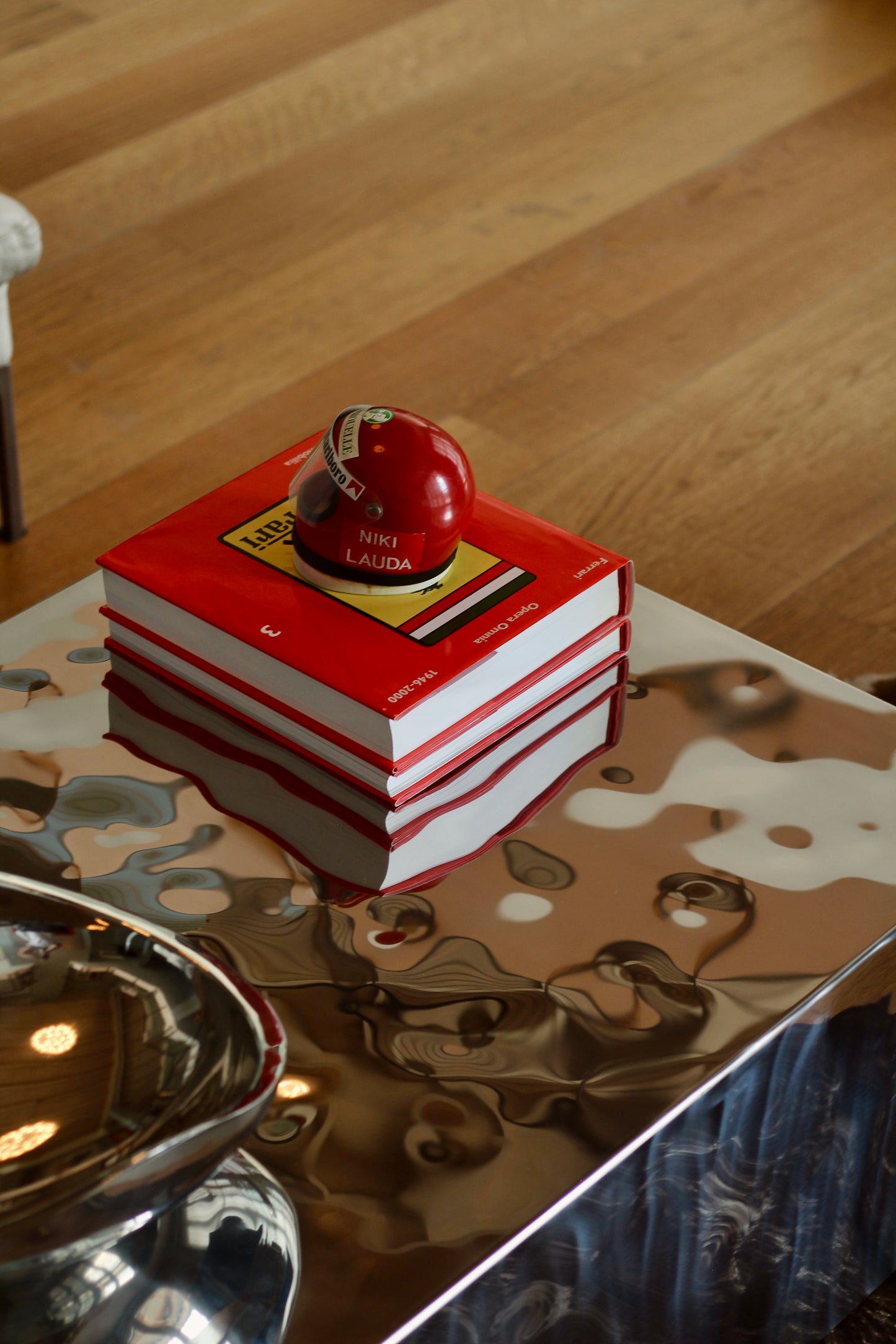 A stack of red and white books with a small red spherical object on top, placed on a reflective metallic table in an indoor setting with a wood floor visible in the background.