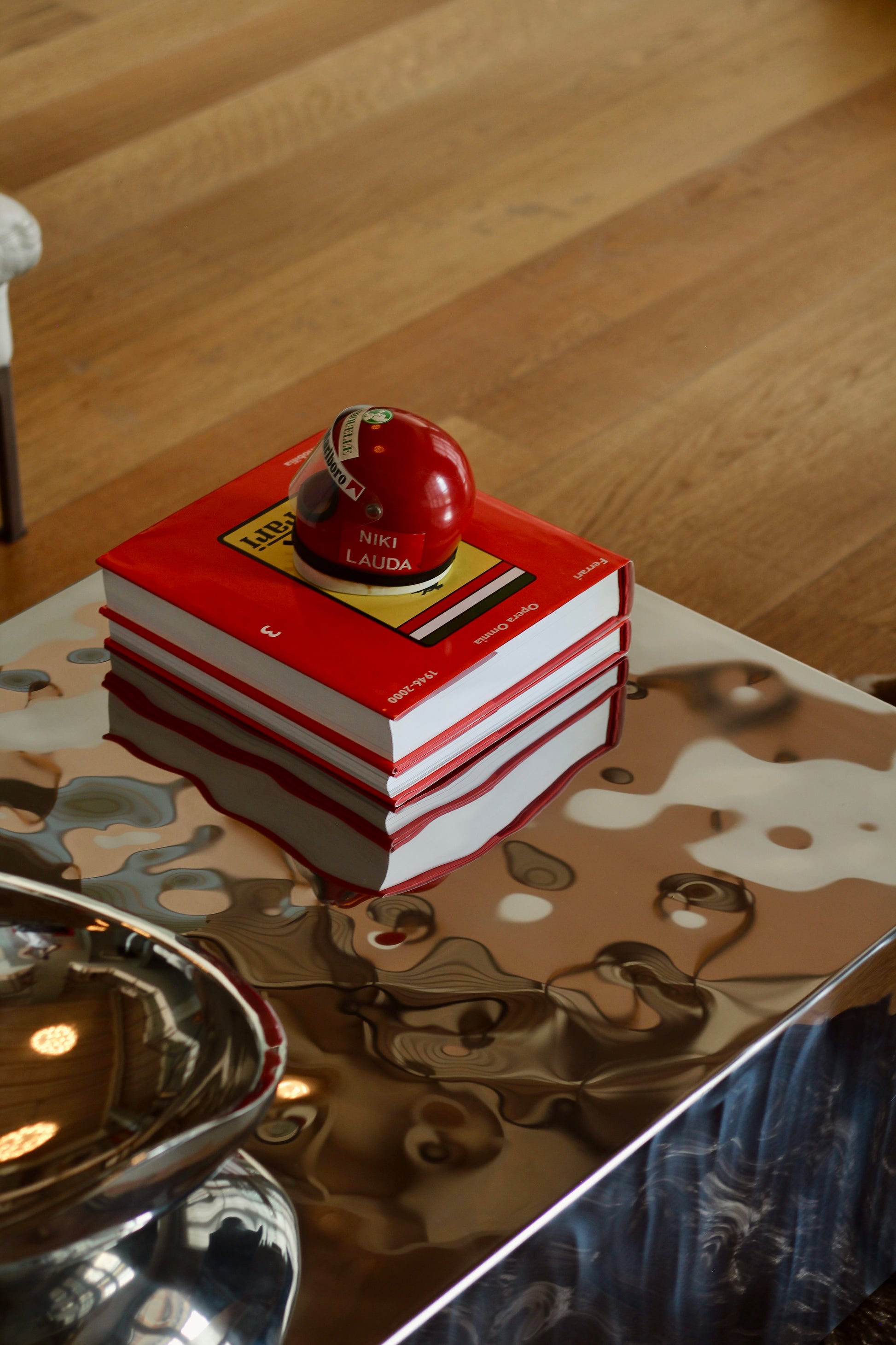 A stack of red and white books with a small red spherical object on top, placed on a reflective metallic table in an indoor setting with a wood floor visible in the background.