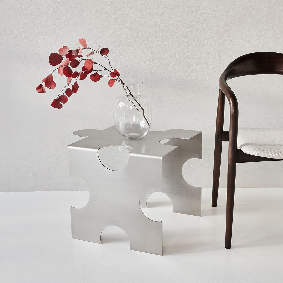 Brushed stainless steel “Puzzle Table” by Veliento styled with a dark wood chair and glass vase with red branches — minimalist contemporary interior in soft natural light.