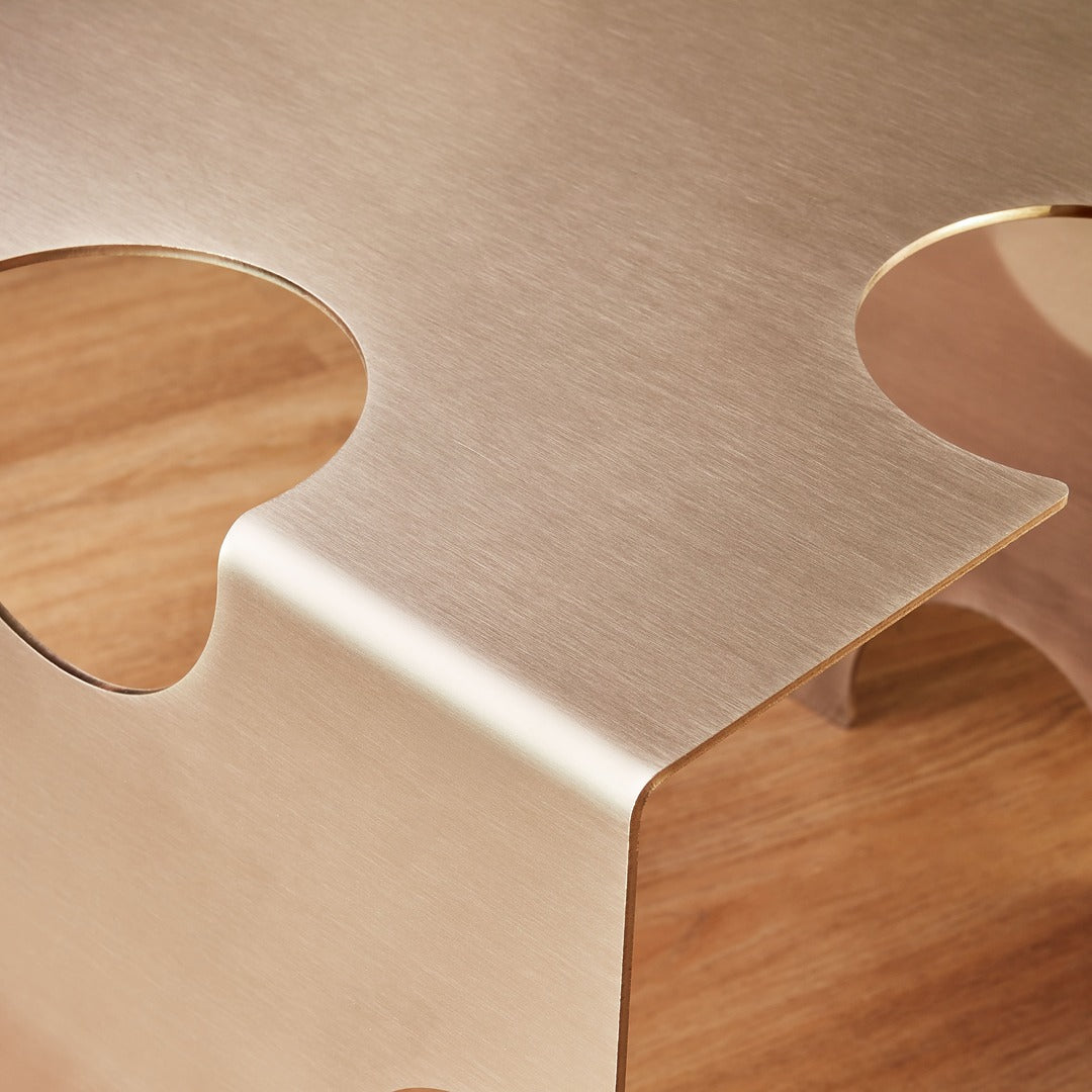 Detail of the “Puzzle Table” by Veliento — brushed stainless steel surface with elegant curves reflecting warm light on a wooden floor.
