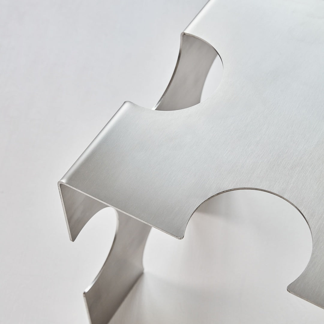 Minimal close-up of the “Puzzle Table” by Veliento — precise brushed stainless steel joints with sculptural contours and seamless bending.