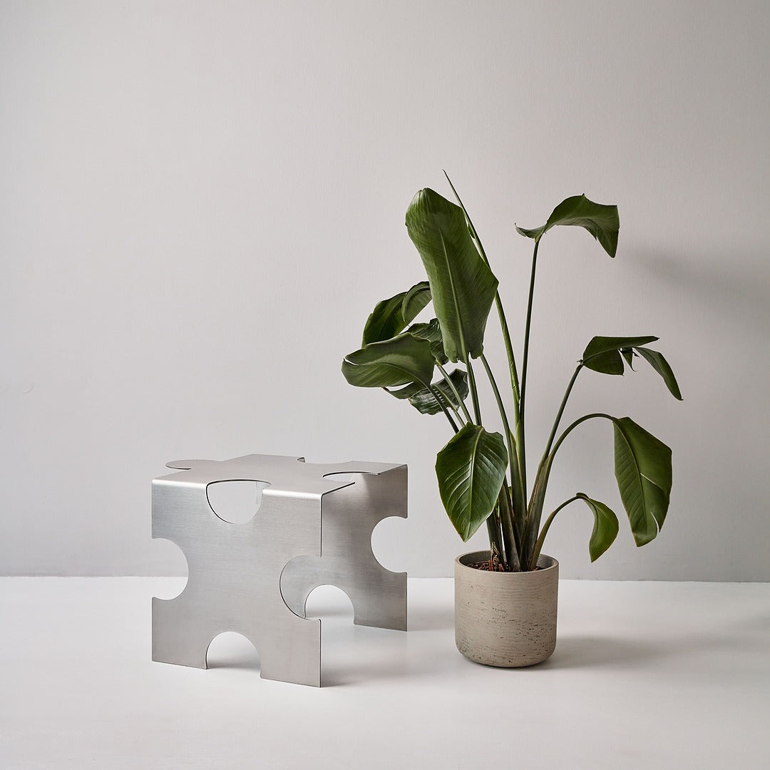 “Puzzle Table” by Veliento — brushed stainless steel side table paired with a potted monstera plant in a minimalist studio setting.