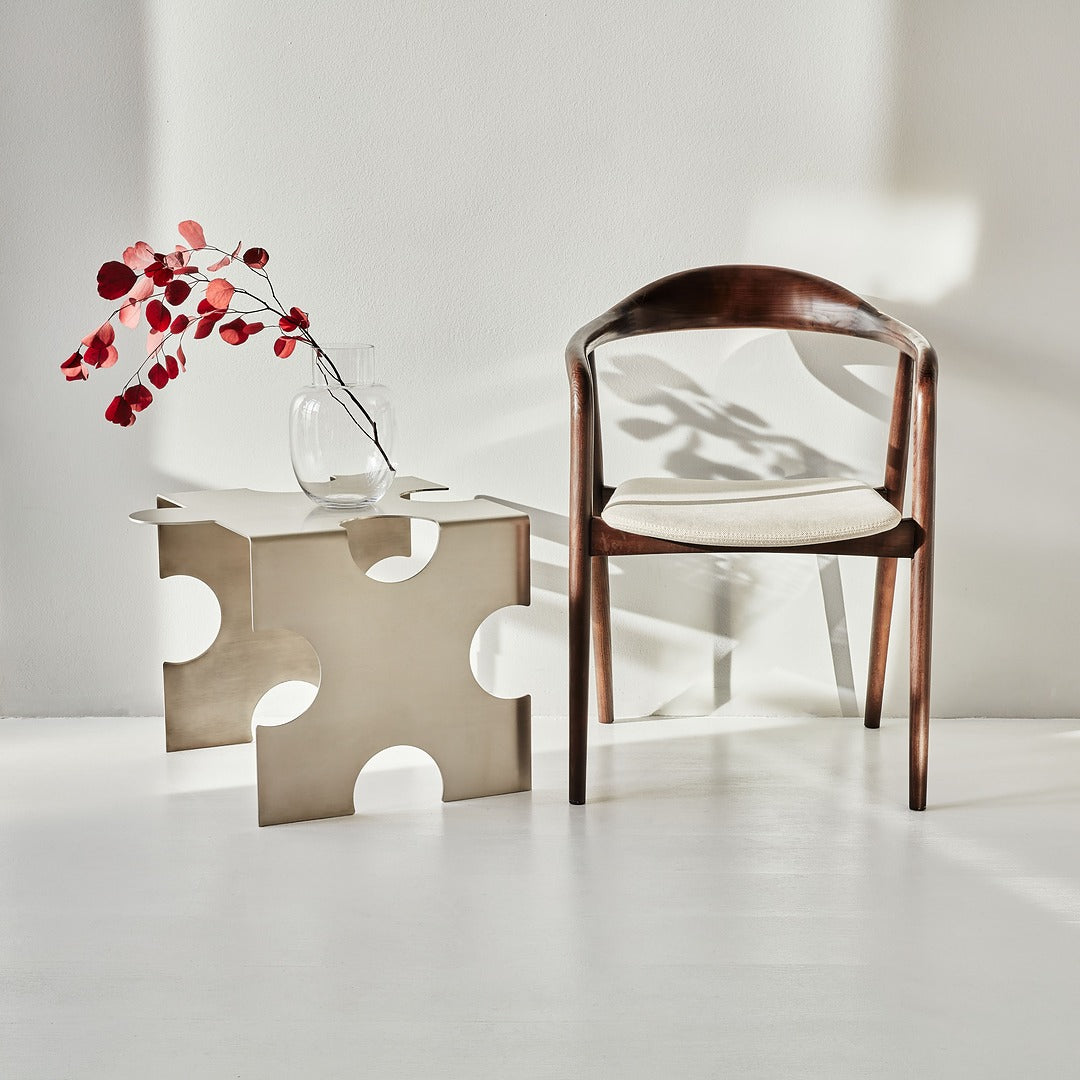 Wooden chair with a white cushion next to a modern side table with a vase and branches on a white floor.